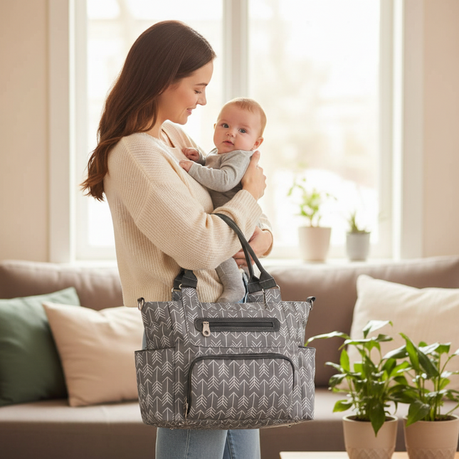 Woman holding a baby and a grey design Mova Mama 6-in-1 convertible diaper tote bag, in a living room.